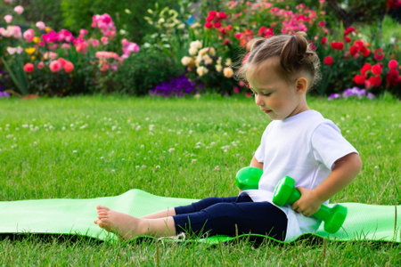 Little cute girl 1-3 in a white t-shirt goes in for sports with dumbbells on a green mat on a background of flowersの写真素材