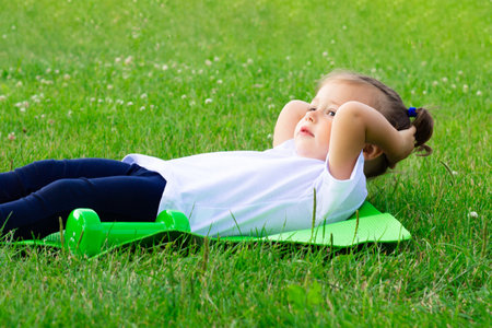 Little cute girl 1-3 in a white t-shirt goes in for sports on a green mat on the grass on a background of flowersの写真素材