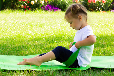 Little cute girl 1-3 in white t-shirt goes in for sports with elastic on green mat on the grass on background of flowersの写真素材