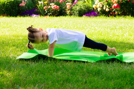 Little cute girl 1-3 in a white t-shirt goes in for sports on a green mat on the grass on a background of flowersの写真素材