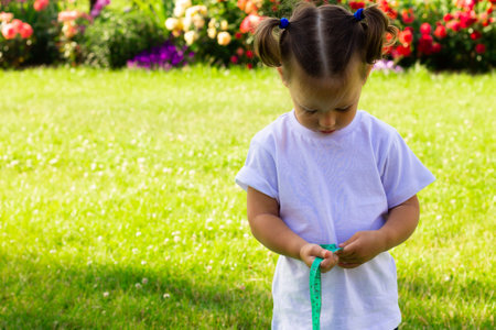Little cute girl 1-3 in white t-shirt measures her waist with a tape after sports on the grass on background of flowersの写真素材