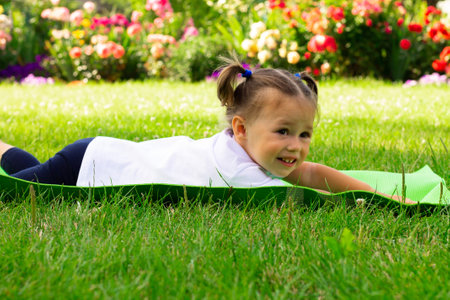Little cute girl 1-3 in a white t-shirt goes in for sports on a green mat on the grass on a background of flowersの写真素材
