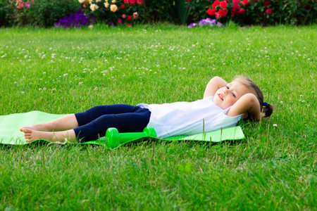Little cute girl 1-3 in a white t-shirt goes in for sports on a green mat on the grass on a background of flowersの写真素材