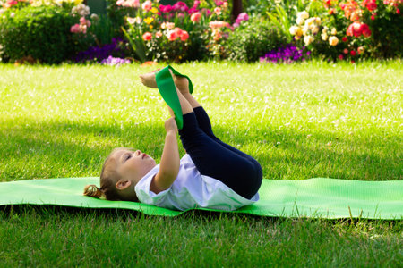 Little cute girl 1-3 in white t-shirt goes in for sports with elastic on green mat on the grass on background of flowersの写真素材