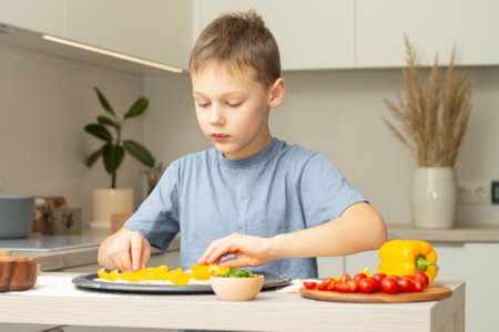 Boy 7-10 in T-shirt cooking pizza in kitchen. Kid arranges ingredients on pizza baseの写真素材