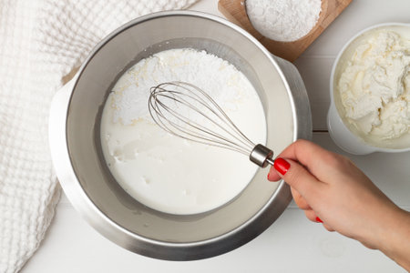 A woman's hand with a whisk over a bowl of whipping cream and powdered sugar near a towel and bowls of powdered sugar and ricotta. Flat lay.の写真素材