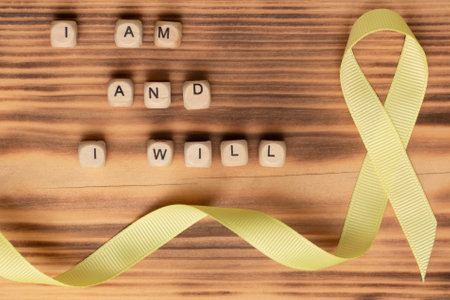 Wooden cubes with text of the slogan of World Cancer Day I am and I will and a yellow ribbon, on a wooden surface. Flat layの写真素材