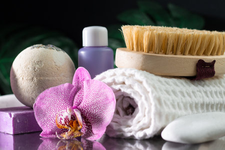 Beautiful lilac orchid flower, cosmetic bottle, massage brush, rolled towel and handmade soap with white stone and monstera leaves on black surface. SPA or beauty salon and procedures conceptの写真素材