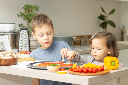 Cute little girl 2-4 in gray dress and boy 7-10 in T-shirt cooking pizza together in kitchen. Brother and sister cookingの写真素材