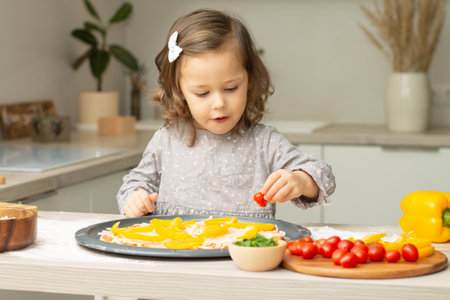 Cute little girl 2-4 in gray dress cooking pizza in kitchen. Kid arranges ingredients on pizza baseの写真素材
