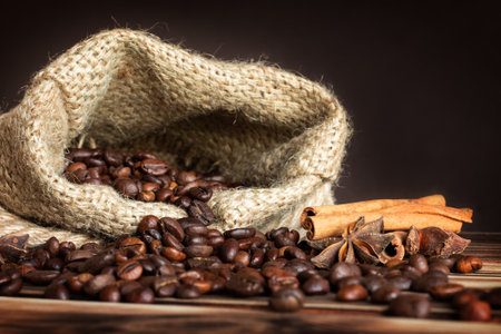 Coarse burlap sack with sprinkled grains of coffee on a burnt wood tabletop with cinnamon sticks and anise stars. Copy spaceの写真素材