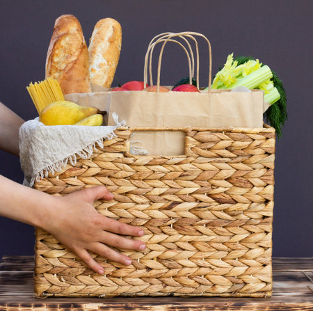 Fresh eco organic vegetables, greens and fruits, cereals and pasta in a wicker basket in the hands of a child. Delivery or donation of ecological farm food conceptの写真素材