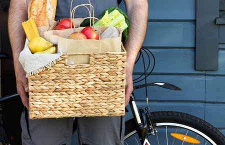 Fresh organic bread, vegetables, greens and fruits, cereals and pasta in a wicker basket in the hands of a bicycle courier. Bike eco-friendly delivery or donation of ecological farm food concept.の写真素材