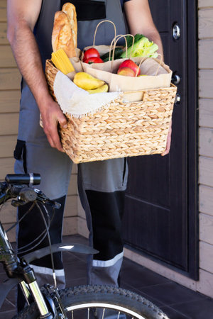 Fresh organic bread, vegetables, greens and fruits, cereals and pasta in a wicker basket in the hands of a bicycle courier. Bike eco-friendly delivery or donation of ecological farm food concept.の写真素材