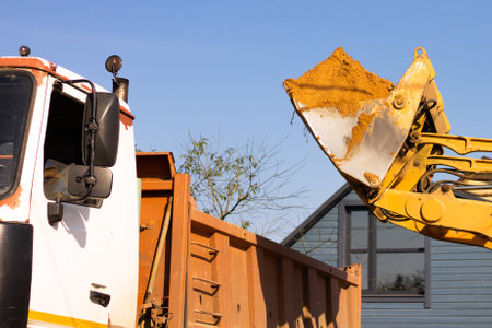A wide metal excavator bucket digs sand or clay, loading it into a dump truck at a construction...