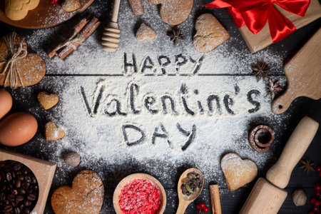 Happy Valentines Day written on flour. Gingerbread heart shaped cookies, spices, coffee beans and baking supplies on black wood backgroundの写真素材