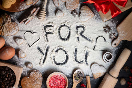 For You written on flour. Gingerbread heart shaped cookies, spices, coffee beans and baking supplies on black wood backgroundの写真素材