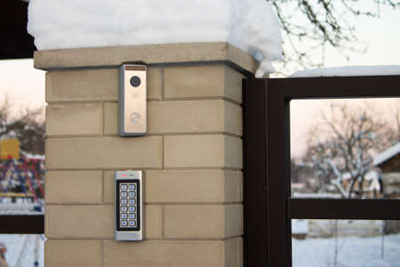 Silver intercom call panel with blue number buttons and a video camera, on a brick beige fence...