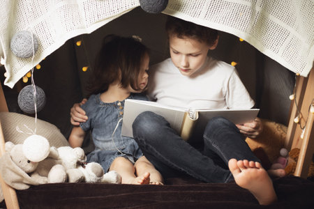 Siblings sit in a hut of chairs and blankets. Brother and sister reading book with flashlight at homeの写真素材