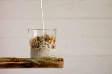 Milk flowing into granola in a transparent glass with rye spike and walnuts on a shelf on white...