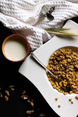 Cereal granola in a white plate with a spoon and a clay glass of milk on a black wooden tabletop....
