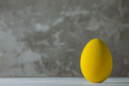 Bright yellow Easter egg, standing on white wooden surface on gray concrete background, copy space