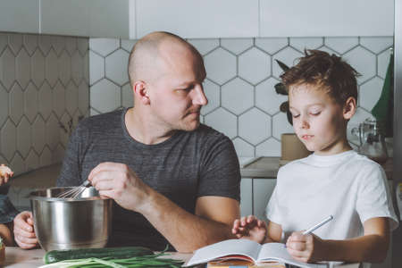 Father whips omelet and helps his son do his homework in the kitchen. Man doing household chores.の写真素材