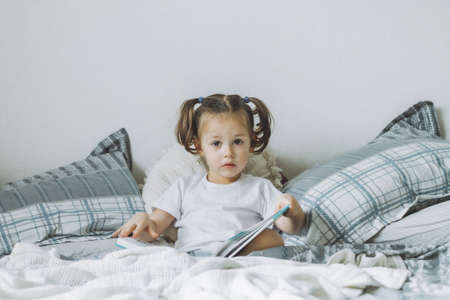 Little dark-haired girl 2-4 with two ponytails sits on bed with pillows reading bookの写真素材