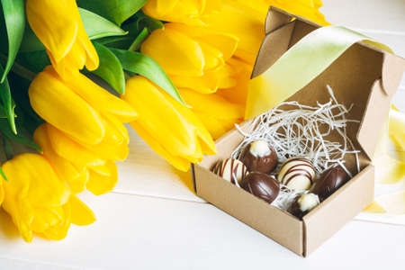 Chocolate candies in craft box and bouquet of yellow tulips, on white wooden surface