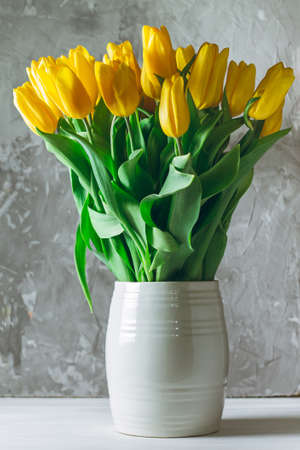 Yellow tulip bouquet in white vase on gray concrete background, verticalの写真素材