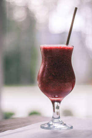 Partially blurred glass of red berry smoothie with straw on wooden table with white tablecloth near window. Verticalの写真素材