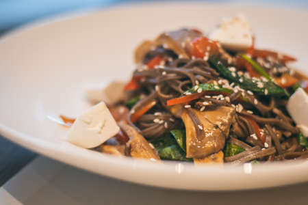 Partially blurred plate of buckwheat noodles with spinach, oyster mushrooms, tomatoes, sesame seeds...