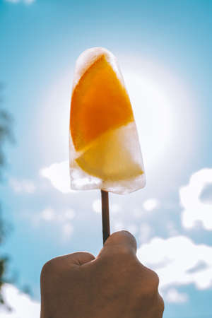 Frozen ice cream on stick in child's hand on background of blue sky. Ice cream with orange and lemon...