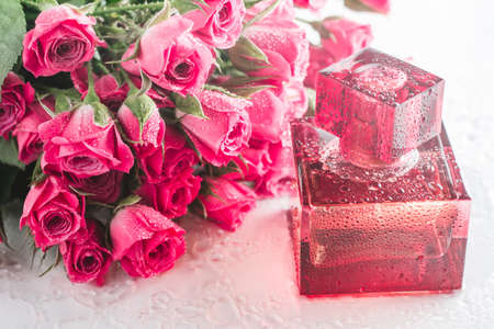 Burgundy square perfume bottle on glossy surface with delicate pink roses and water drops. Eau de...