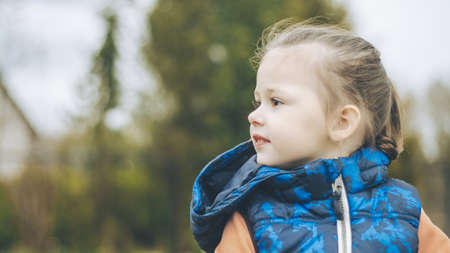 Child in blue vest looks away and smiles. Girl 3-4 on background of blurry treesの写真素材