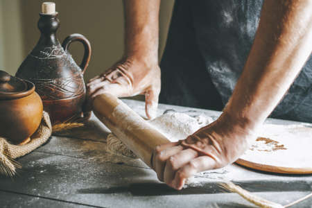 Male hands roll dough and flour with rolling pin next to clay pot and oil bottle on dark table while...