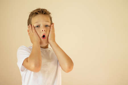 A surprised or scared boy of 7-10 years old in a white T-shirt stands and shouts with his hands on his cheeks on a beige background. Copy space.の写真素材