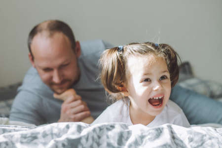 Father plays with little daughter 2-4 on bed. Dad tickles kids feet with his beard. Family having...