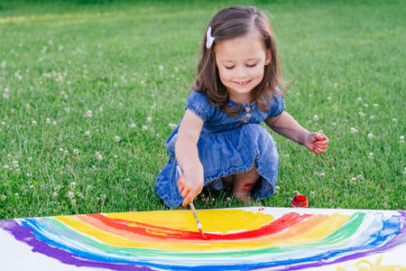 Little girl 2-4 years old paints rainbow and sun on large sheet of paper sitting on green lawnの写真素材