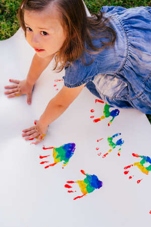 Little girl 2-4 draws leaving rainbow colors child's handprints on large white sheet of paper. Verticalの写真素材