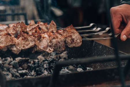 Male hand turns raw fried meat on skewers is cooked on charcoal grill. Cooking barbecue outdoorsの写真素材