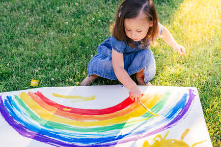 Little girl 2-4 years old paints rainbow and sun on large sheet of paper, sitting on green lawn in sunlightの写真素材