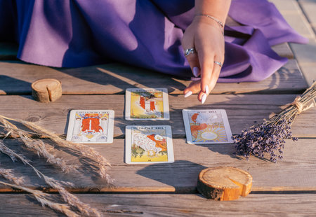 Woman's hand with purple nails points to four Tarot cards spread out on wooden surface next to spikelets and lavender. Minsk, Belarus - 07.28.2021のeditorial素材