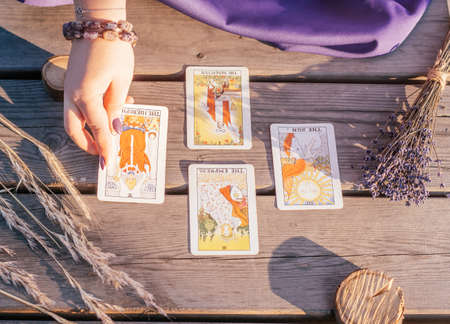 Woman's hand with purple nails lays out one of four Tarot cards spread out on wooden surface next to spikelets and lavender. Minsk, Belarus - 07.28.2021のeditorial素材