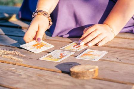 Woman's hand with purple nails lays out one of four Tarot cards spread out on wooden surface next to...