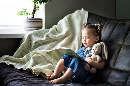 Child reading. Little cute girl 1-3 is reading a book with a toy plush hare sitting on a sofa