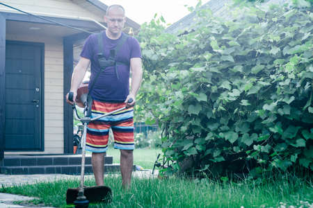 Man in goggles, shorts and T-shirt mows lawn with gasoline trimmer against background of vineyard...