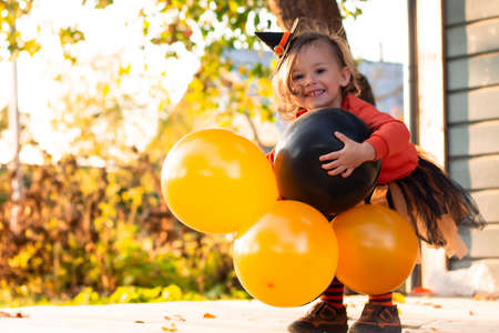A cute little girl 2-3 in an orange and black Halloween witch costume with balloons, standing on the terrace of a wooden gray houseの写真素材