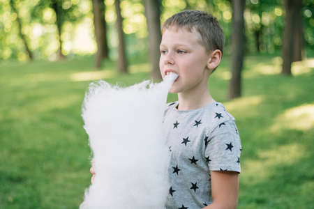 Boy 7-10 eating cotton candy in sunny park among tall trees on green grassの写真素材
