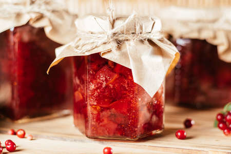 Jars of homemade lingonberry and pear jam with craft paper on lids on wooden surface next to fresh...